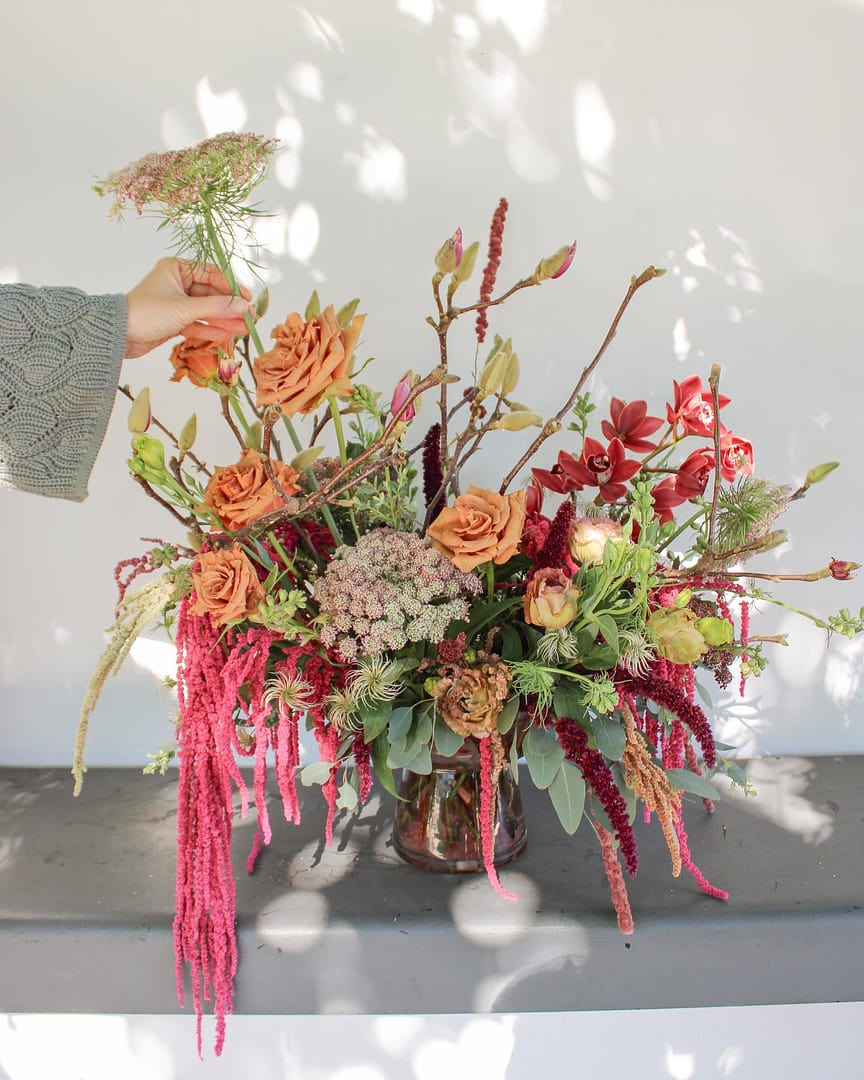 A hand is adjusting a floral arrangement in a glass vase, featuring orange roses, red amaranth draping, and various greenery. The bouquet is placed on a grey surface with a shadow pattern cast upon it, suggesting a sunny environment.